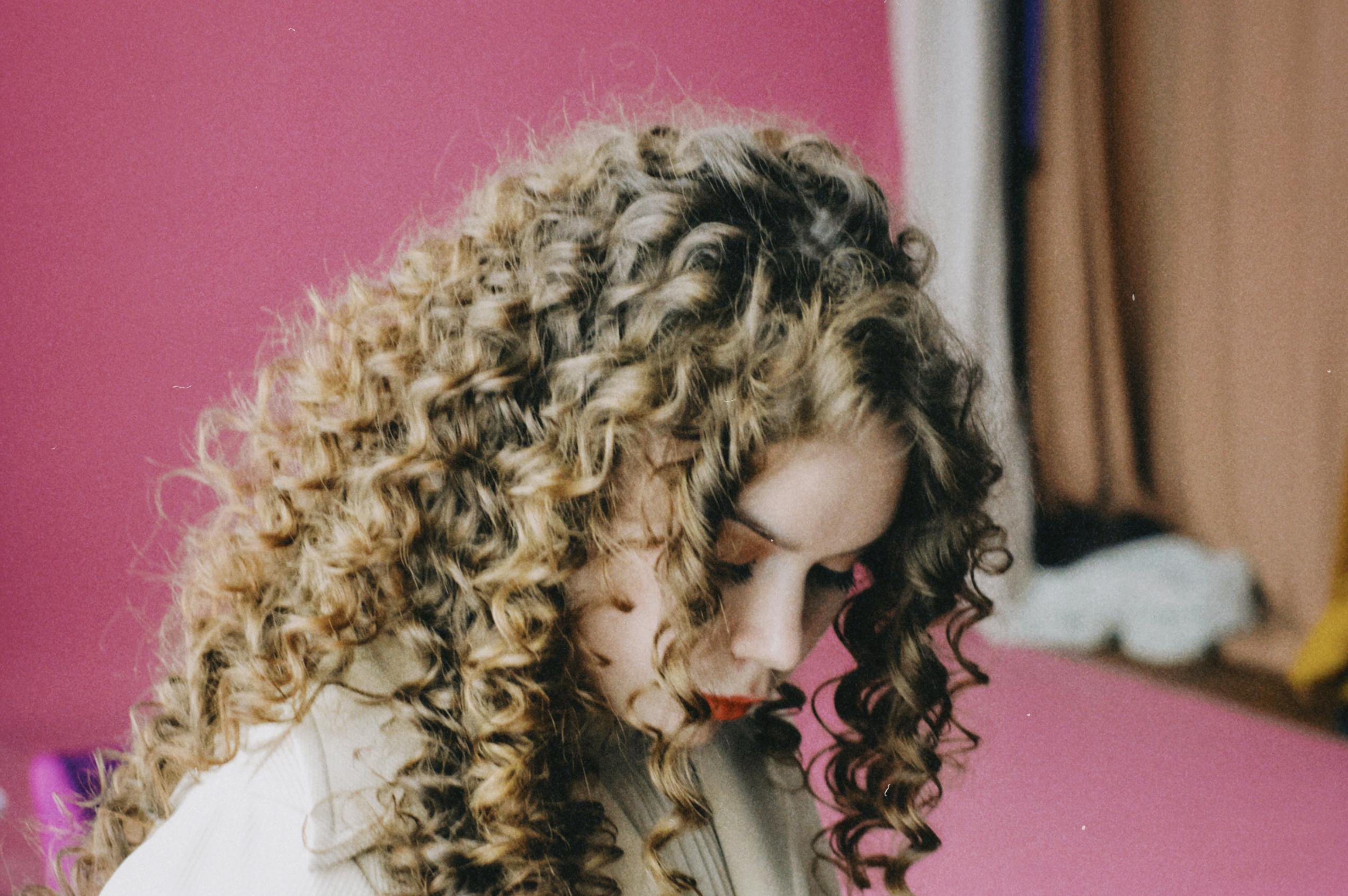 Artistic portrait of a woman with curly hair looking down against a pink background.