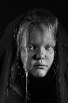 Intense black and white portrait of a teenager with a somber expression, cloaked in darkness.