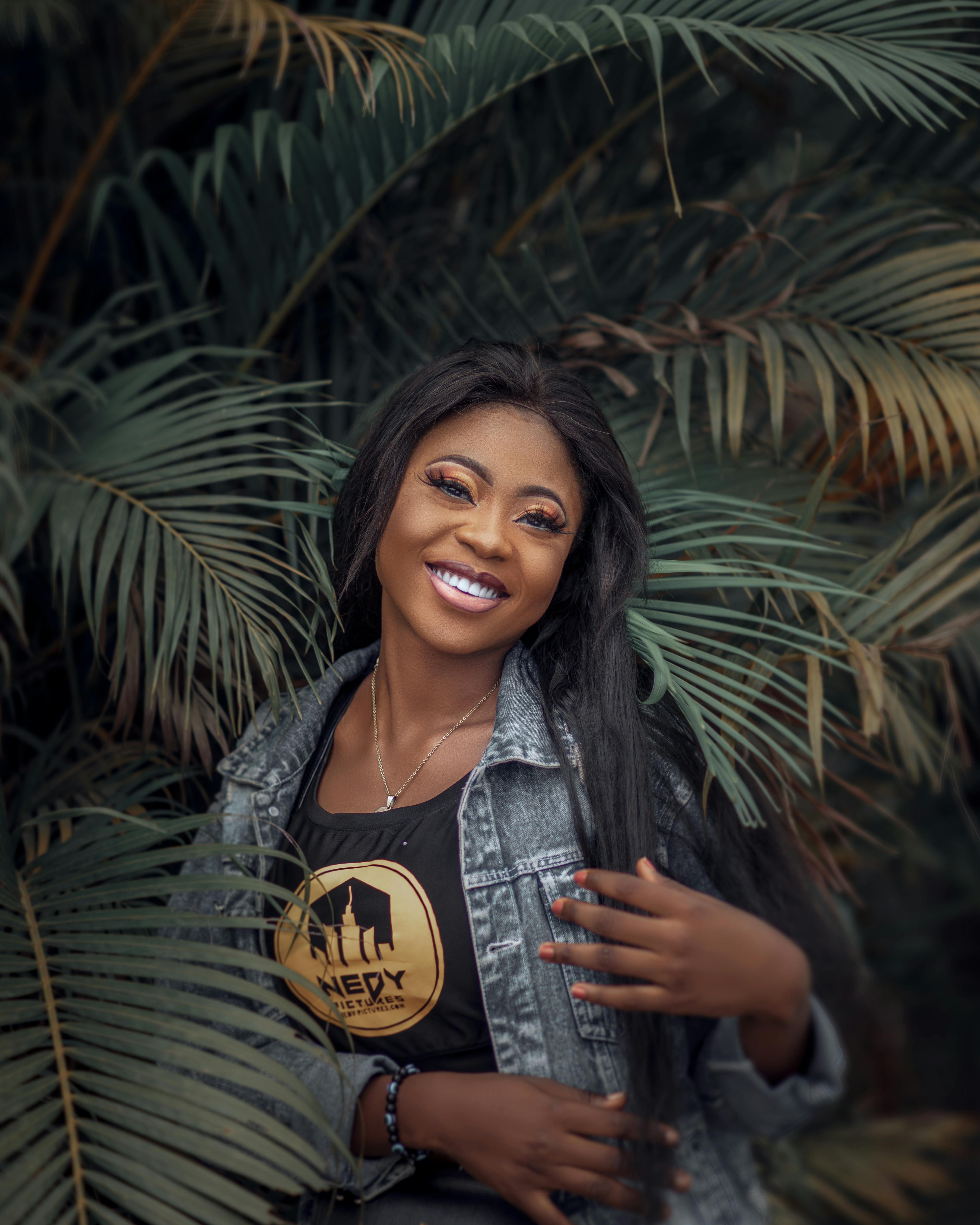 A joyful woman in Lagos posing playfully in a denim jacket surrounded by palm leaves.