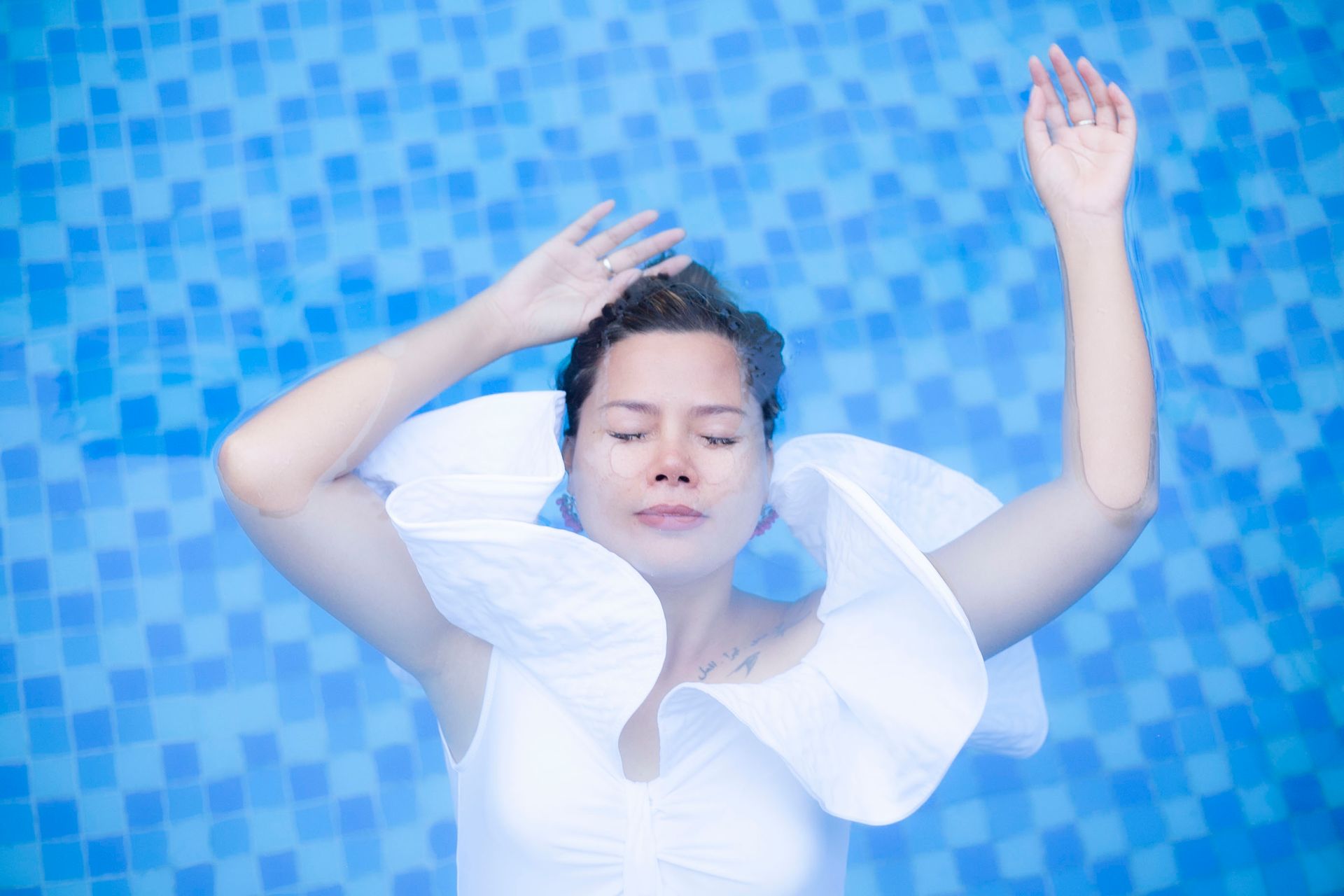 A serene woman in a white dress floats gracefully in a swimming pool with eyes closed.