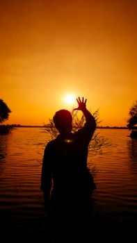 Silhouette of a person raising hand against an orange sunset over a calm lake in India.