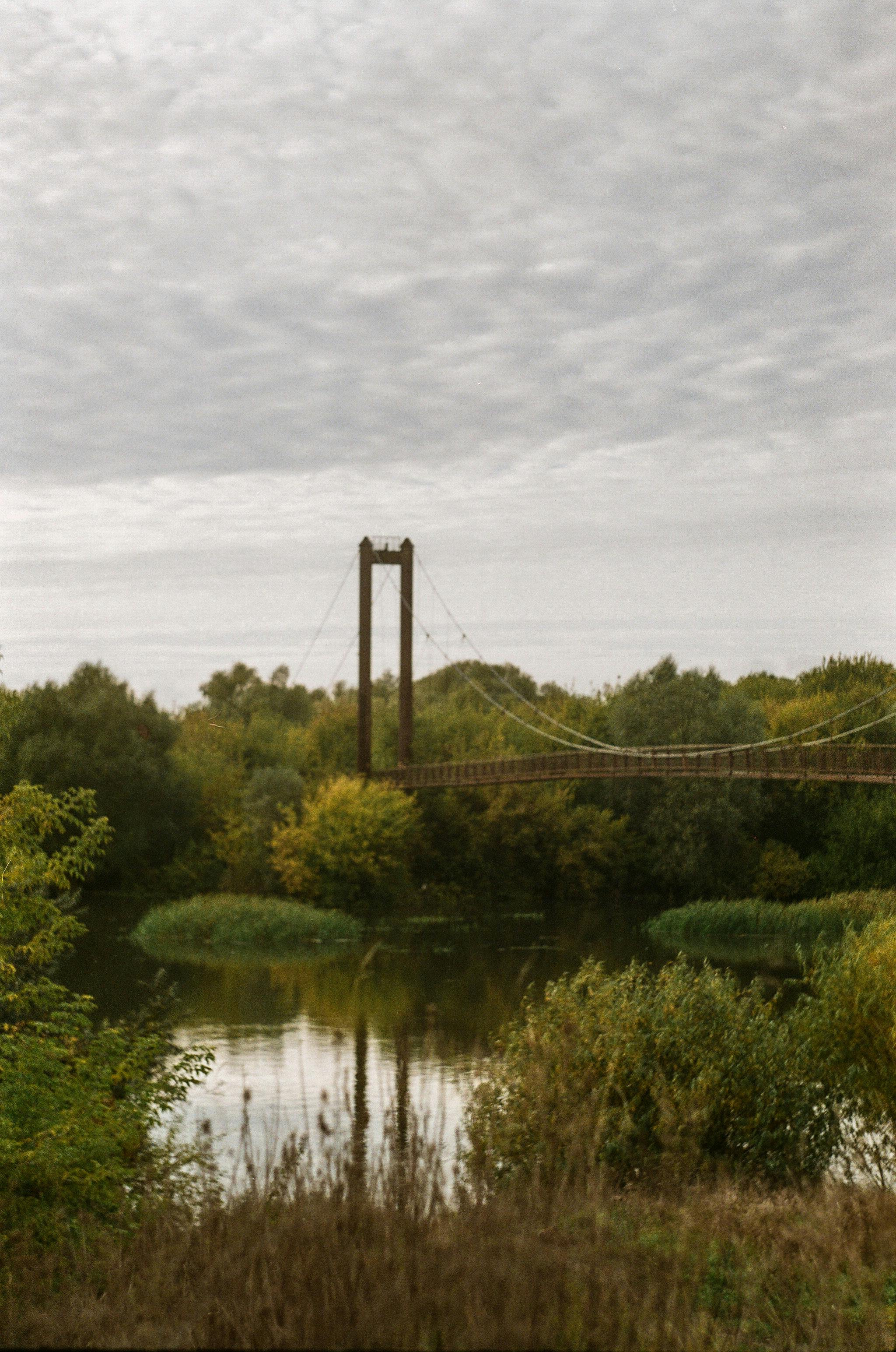 A suspension bridge spans over a serene river surrounded by lush autumn foliage under an overcast sky.