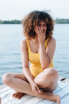 Curly-haired woman in a yellow swimsuit enjoying a sunny day by the lake.