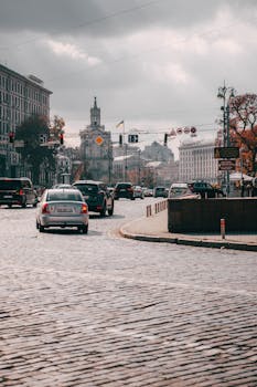 City traffic on a cobblestone street under a cloudy sky, showcasing urban architecture.