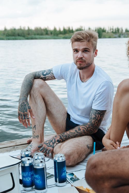 Free A tattooed man in a white t-shirt sits on a pier by the lake, enjoying a summer day. Stock Photo