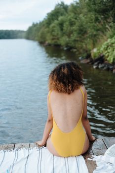 A woman in a yellow swimsuit sits on a pier, enjoying a serene lakeside view.