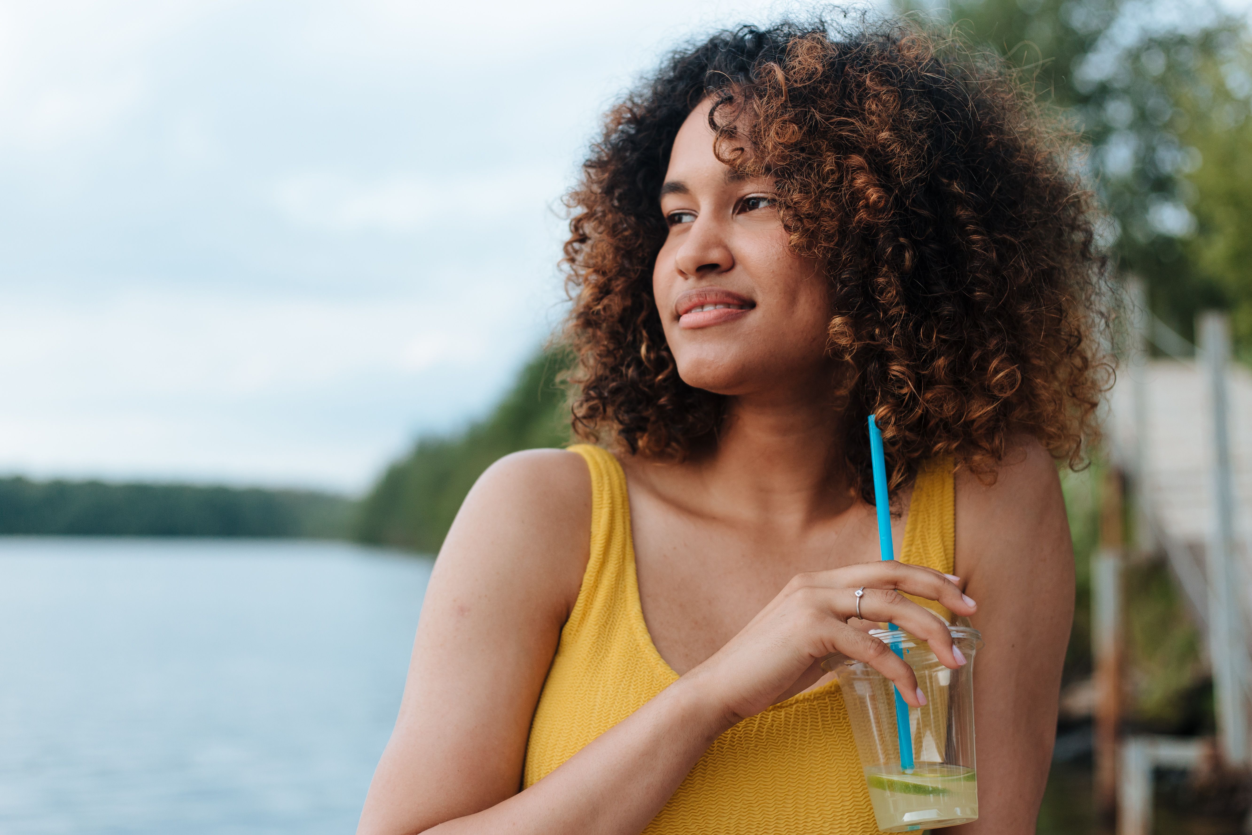 Woman in yellow top sipping a drink by a lake, exuding relaxation and joy in summer.