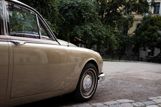 一辆老式米色汽车停在鹅卵石铺就的街道上，车身映着附近树木的倒影