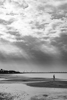 Monochrome beach scene featuring a lone figure under dramatic skies, capturing solitude and nature's vastness.