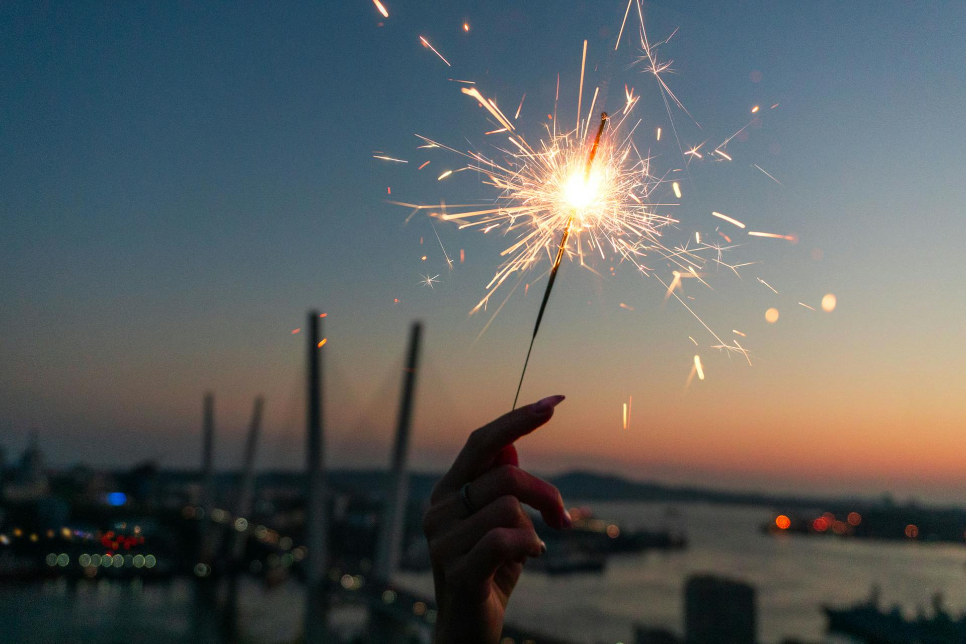 Hand holding sparkler at sunset with a view of Vladivostok bridge and harbor, creating a festive atmosphere.