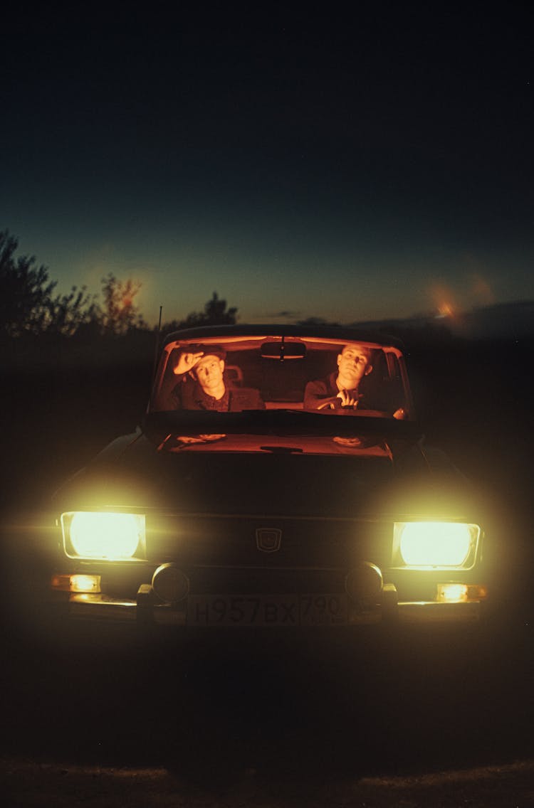 Front View Of A Two Males Sitting In Car With Lights Turned On And During Night Time 