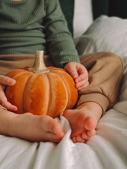 Young child cradles a small pumpkin during fall indoors, capturing autumn coziness.