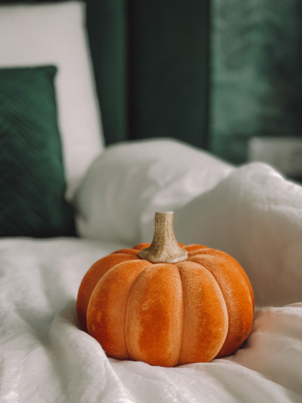 Small orange velvet pumpkin resting on white bedding with green pillows