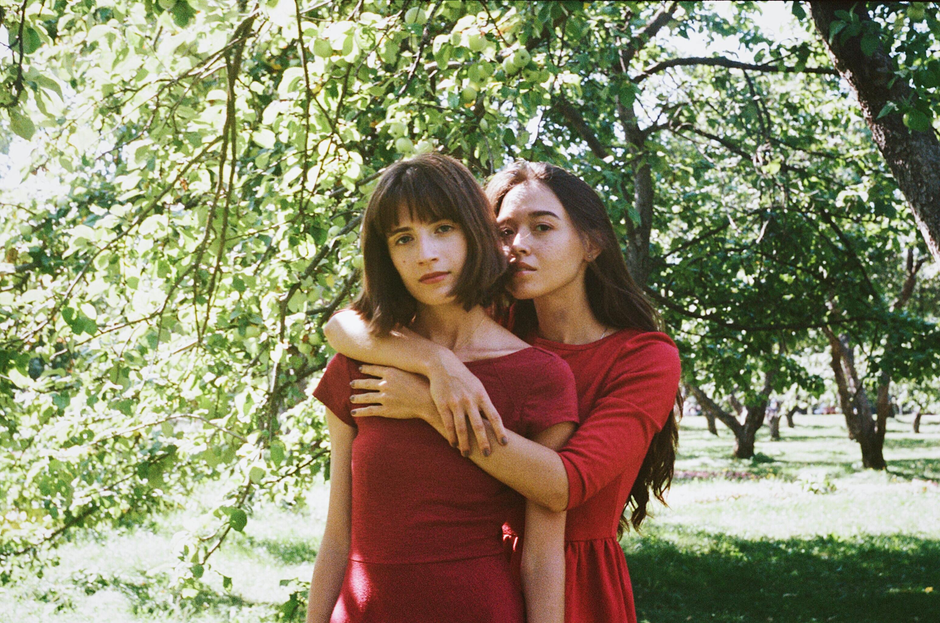 Two women in red dresses embracing in a sunlit apple orchard. Warm and serene.