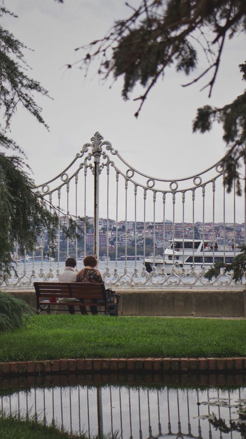 Free Couple sitting on a park bench by a waterfront with a ferry passing by, framed by ornate metal fence. Stock Photo