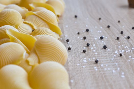 A close-up image of uncooked pasta shells with black pepper and salt on a wooden surface.