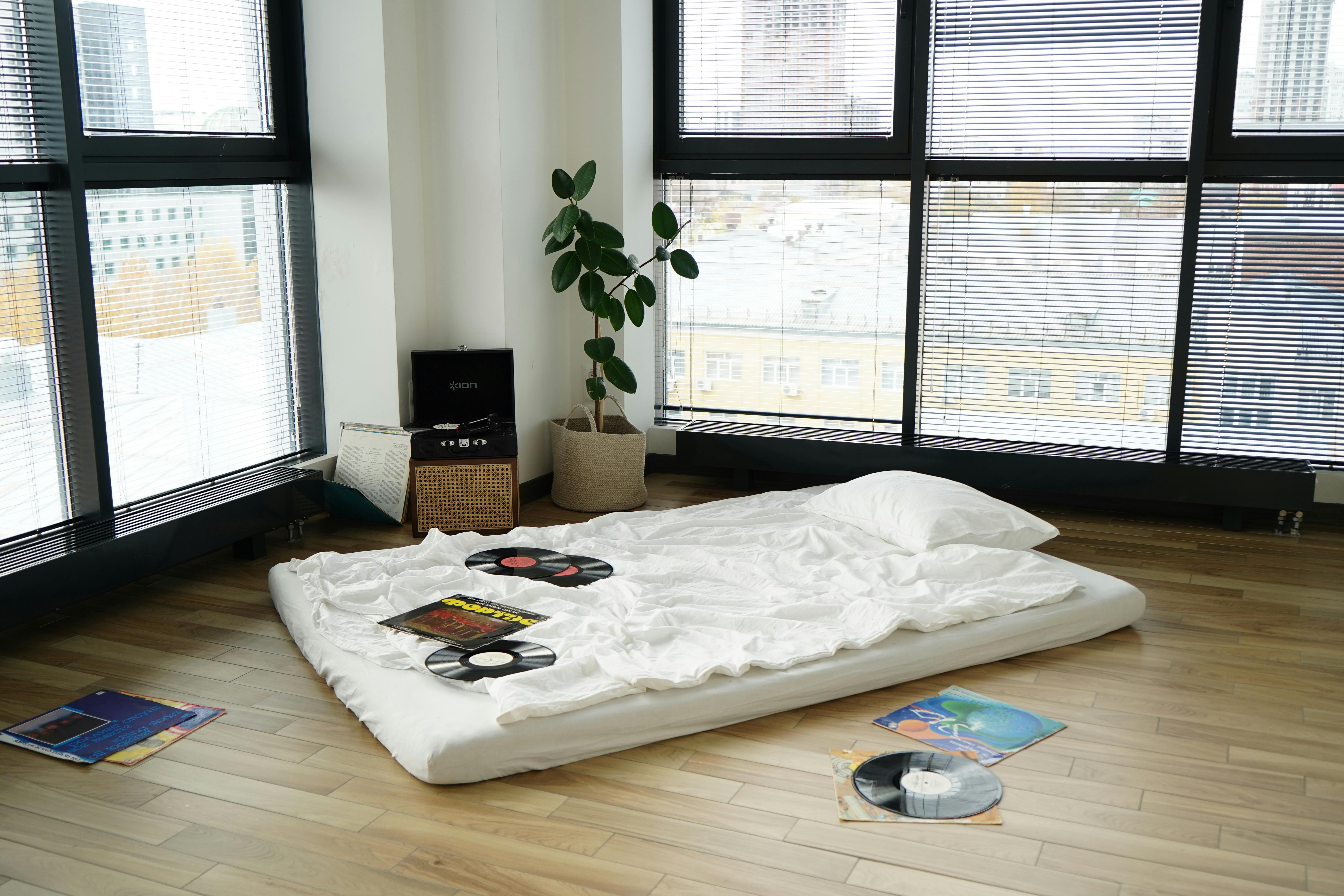 A minimalist apartment interior with a mattress on the floor surrounded by vinyl records and a record player.