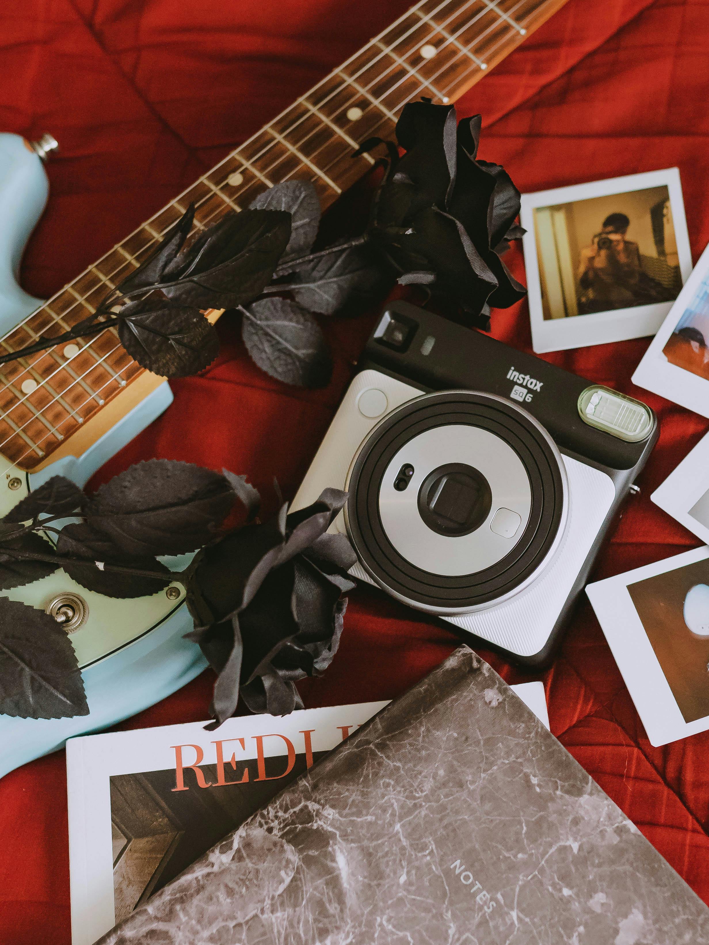Artistic flat lay of a vintage Instax camera, black roses, polaroid photos, and a guitar on a red surface.