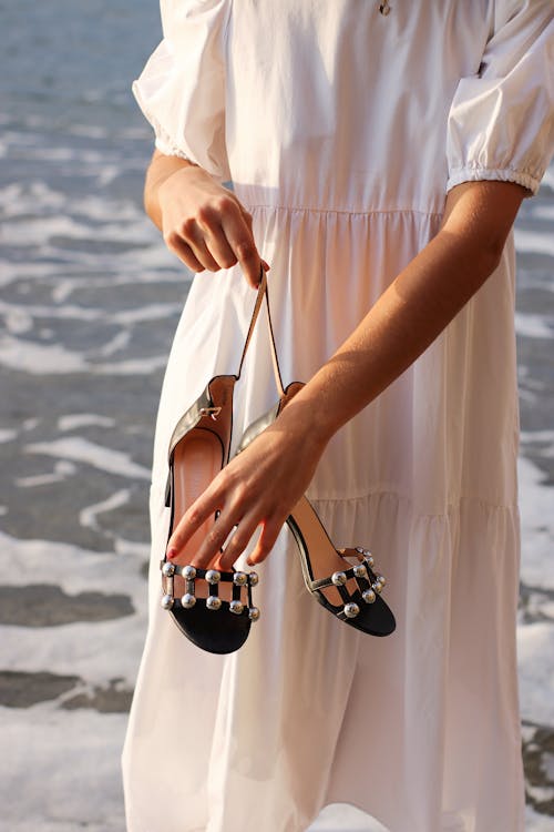 Person in White Dress Holding Shoes with Water in Background