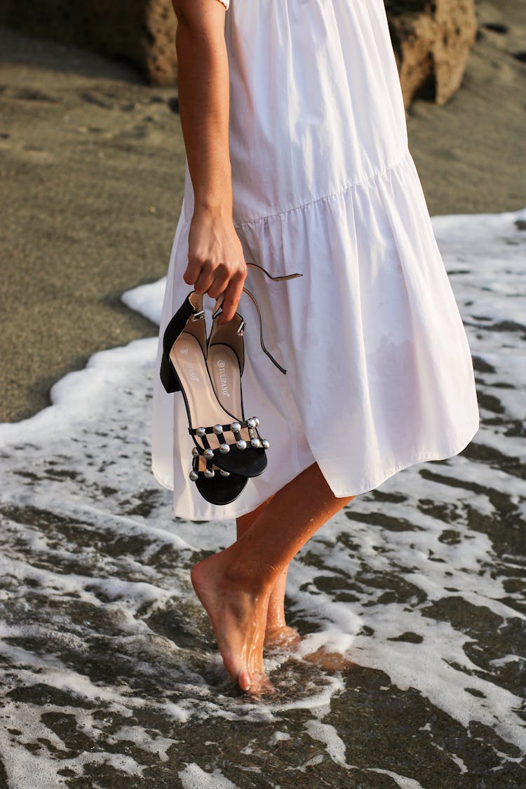 Person In White Dress Holding Shoes And Walking Into Water 