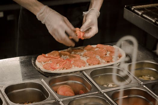 Chef wearing gloves prepares a gourmet pizza with fresh toppings in a professional kitchen.