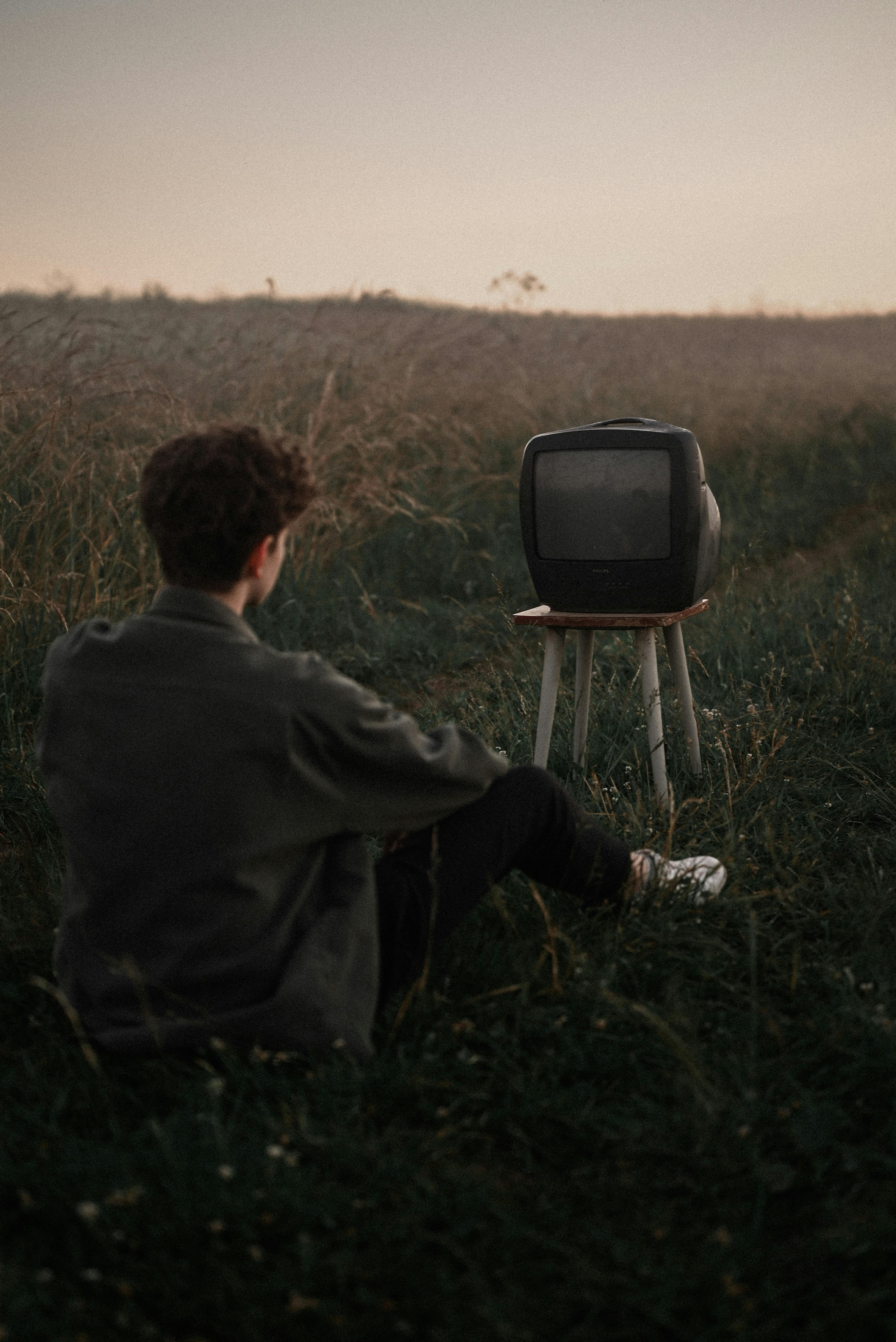 A man sits in a field watching an old TV at dusk, offering a nostalgic and surreal scene.