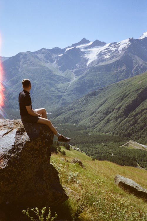 Free A Person Sitting on a Rock Formation Overlooking a Snow Capped Mountain Stock Photo