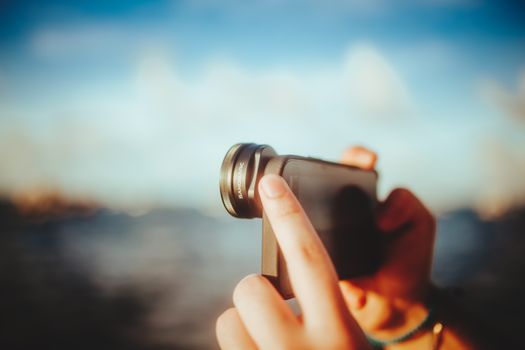 A close-up of hands using a smartphone with a lens to capture outdoor images, against a blurred background.