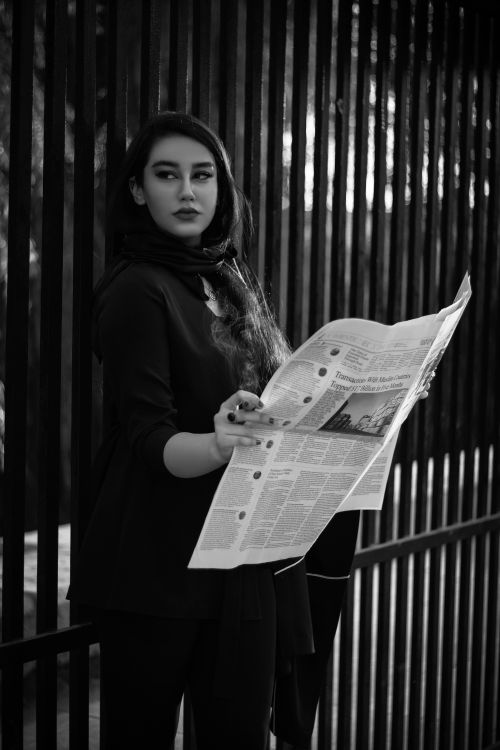 Free A woman in elegant attire reads a newspaper beside an iron fence, captured in black and white. Stock Photo
