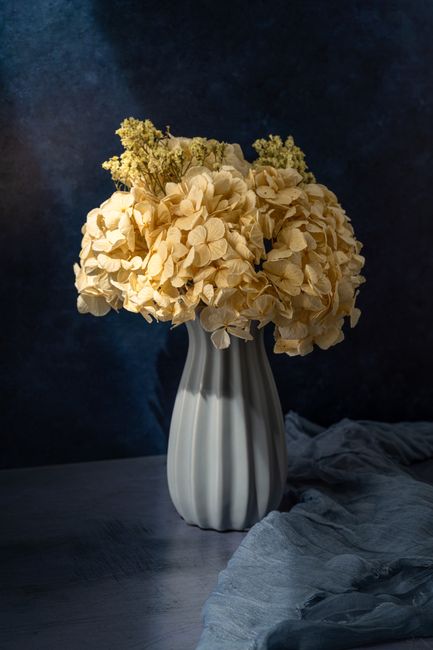 A classic still life of yellow flowers in a ceramic vase on a table against a dark backdrop.