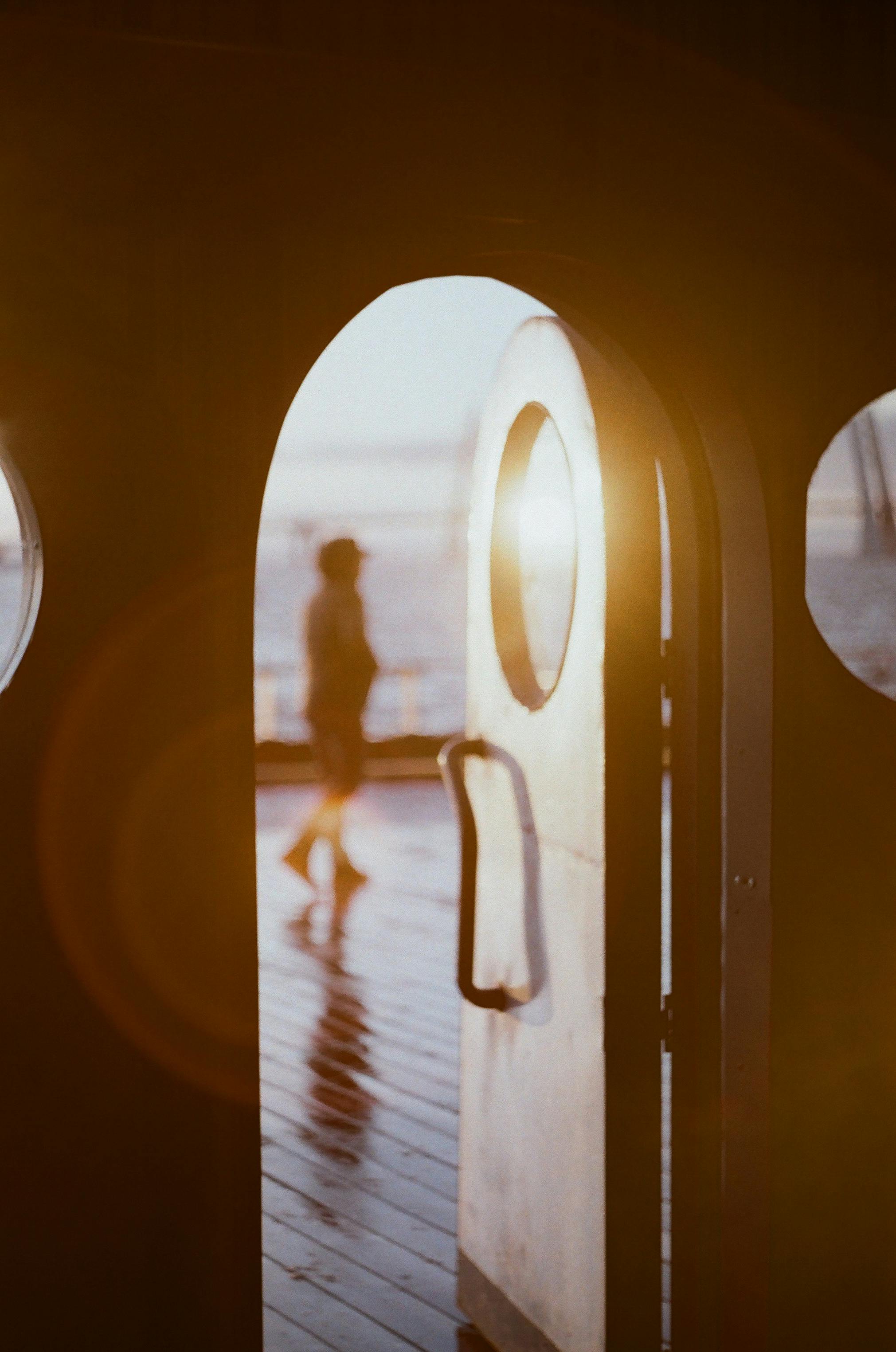 A blurred silhouette is seen through an open sunlit doorway with warm tones.