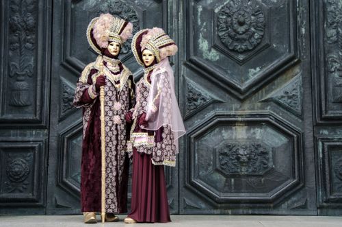 Free A man and woman in elaborate Venetian costumes pose by an ornate door, highlighting festival elegance. Stock Photo