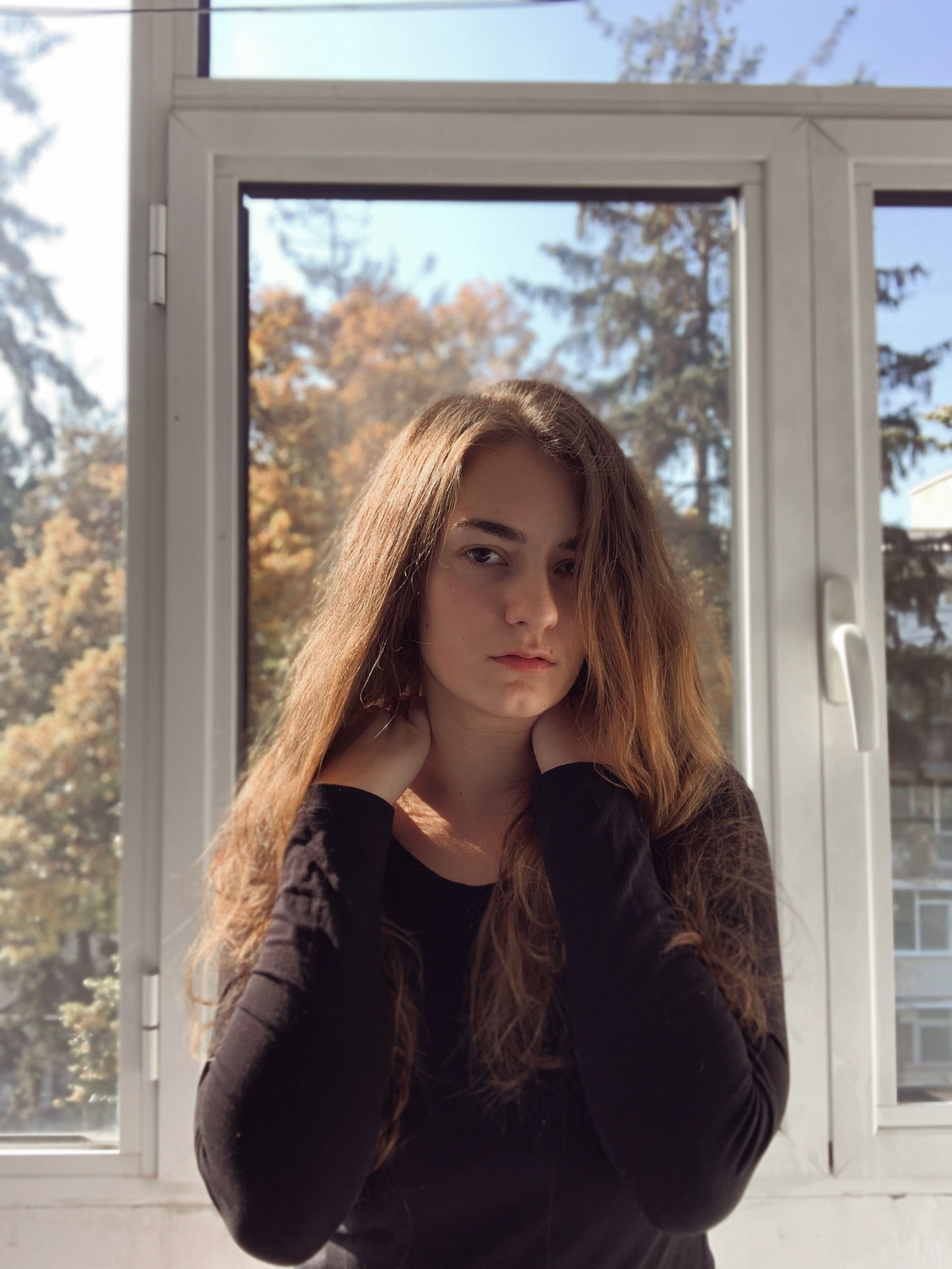 A brunette woman in black long sleeves poses by a sunlit window with a fall background.