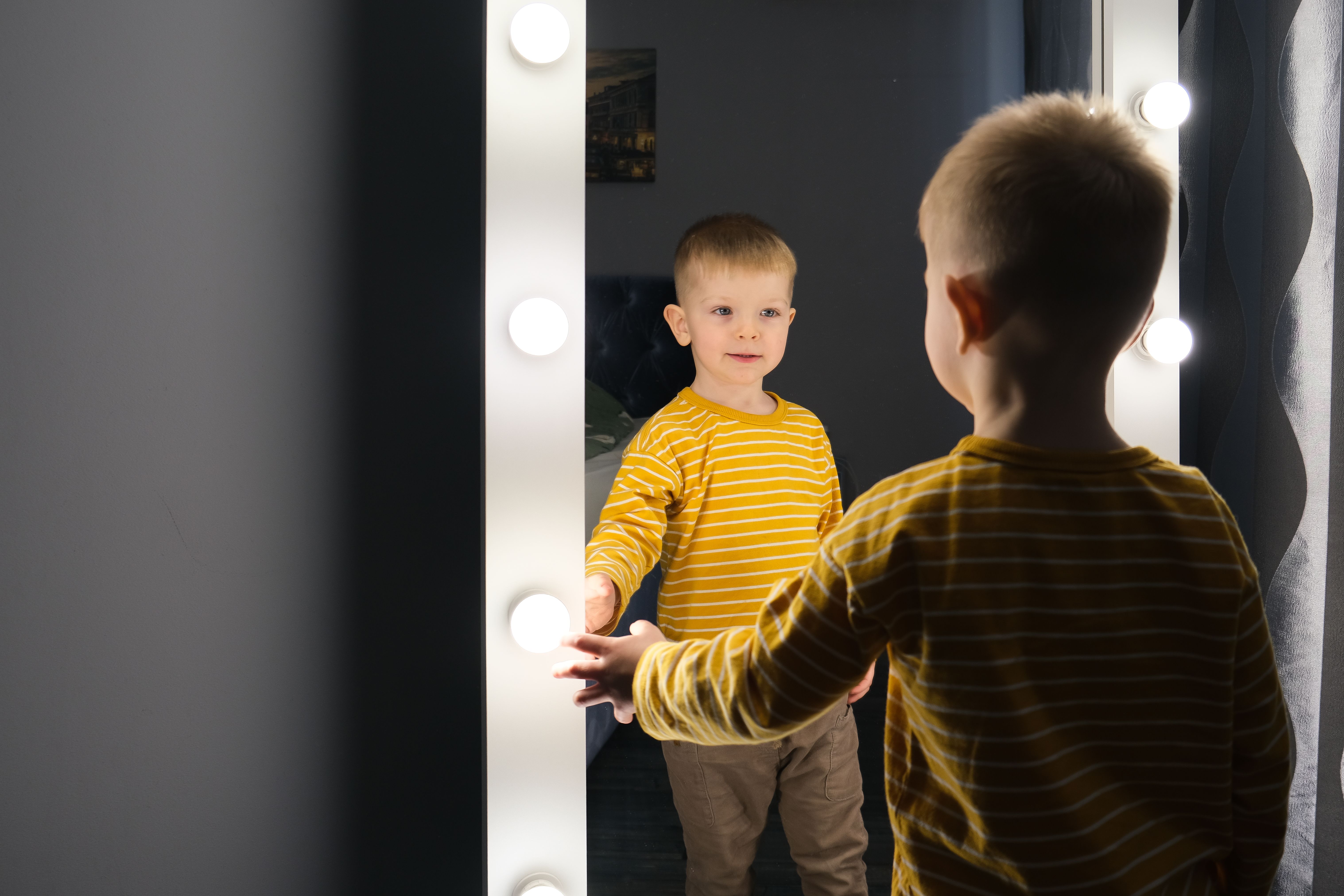 Young boy in a striped shirt looking at his reflection in a mirror surrounded by lights.