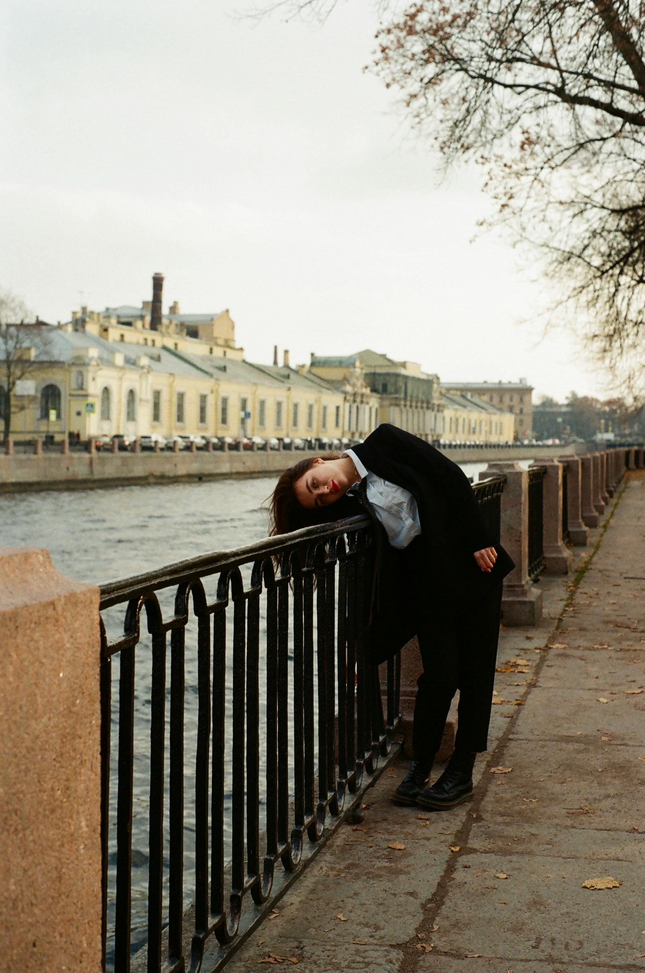 A person in a relaxed pose leaning on a handrail by a river promenade on a cloudy day.