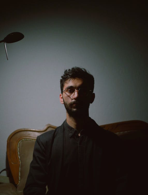 Bearded Young Man in Specs Sitting on Sofa in Dark Room · Free Stock Photo