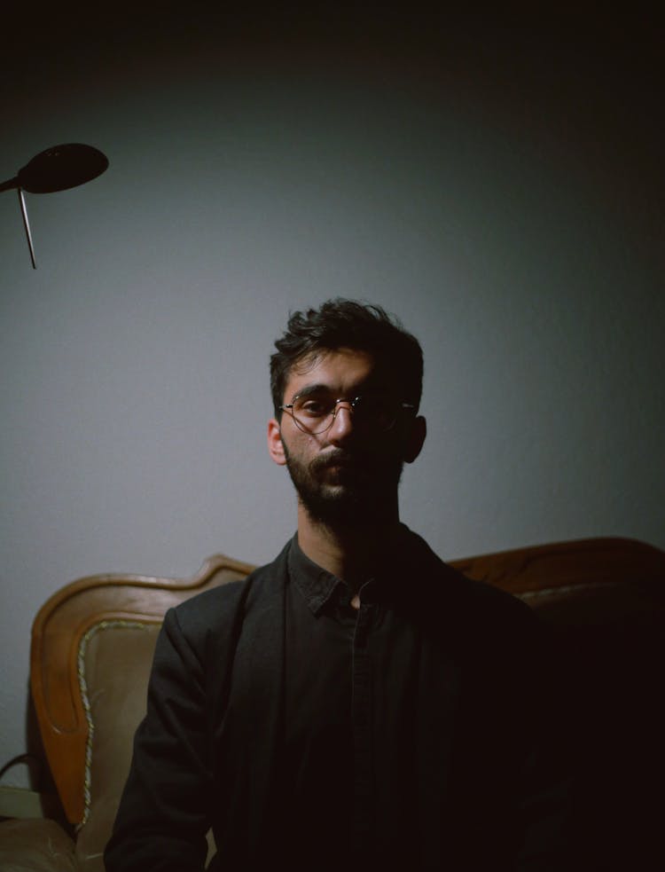 Bearded Young Man In Specs Sitting On Sofa In Dark Room