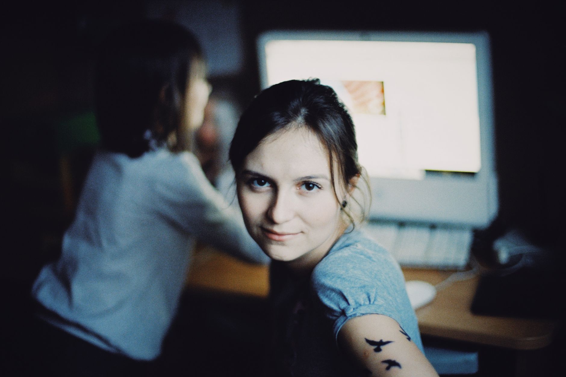A young woman with a bird tattoo smiles at the camera, with a computer in the background.