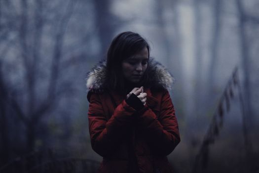 A solitary woman stands clasping her hands in a misty, winter forest.