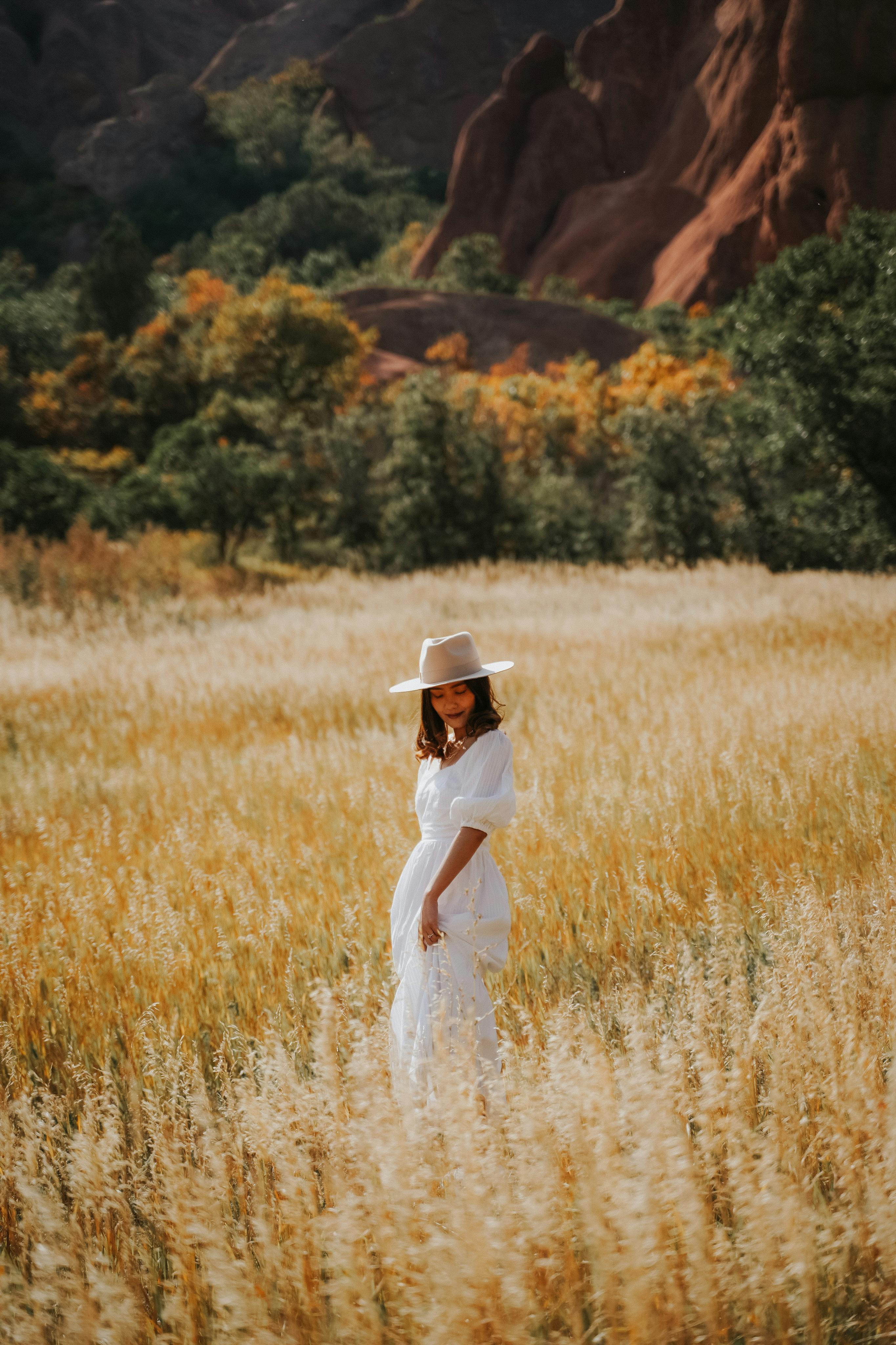 A woman in a white dress and hat smiles in a scenic sunlit field.