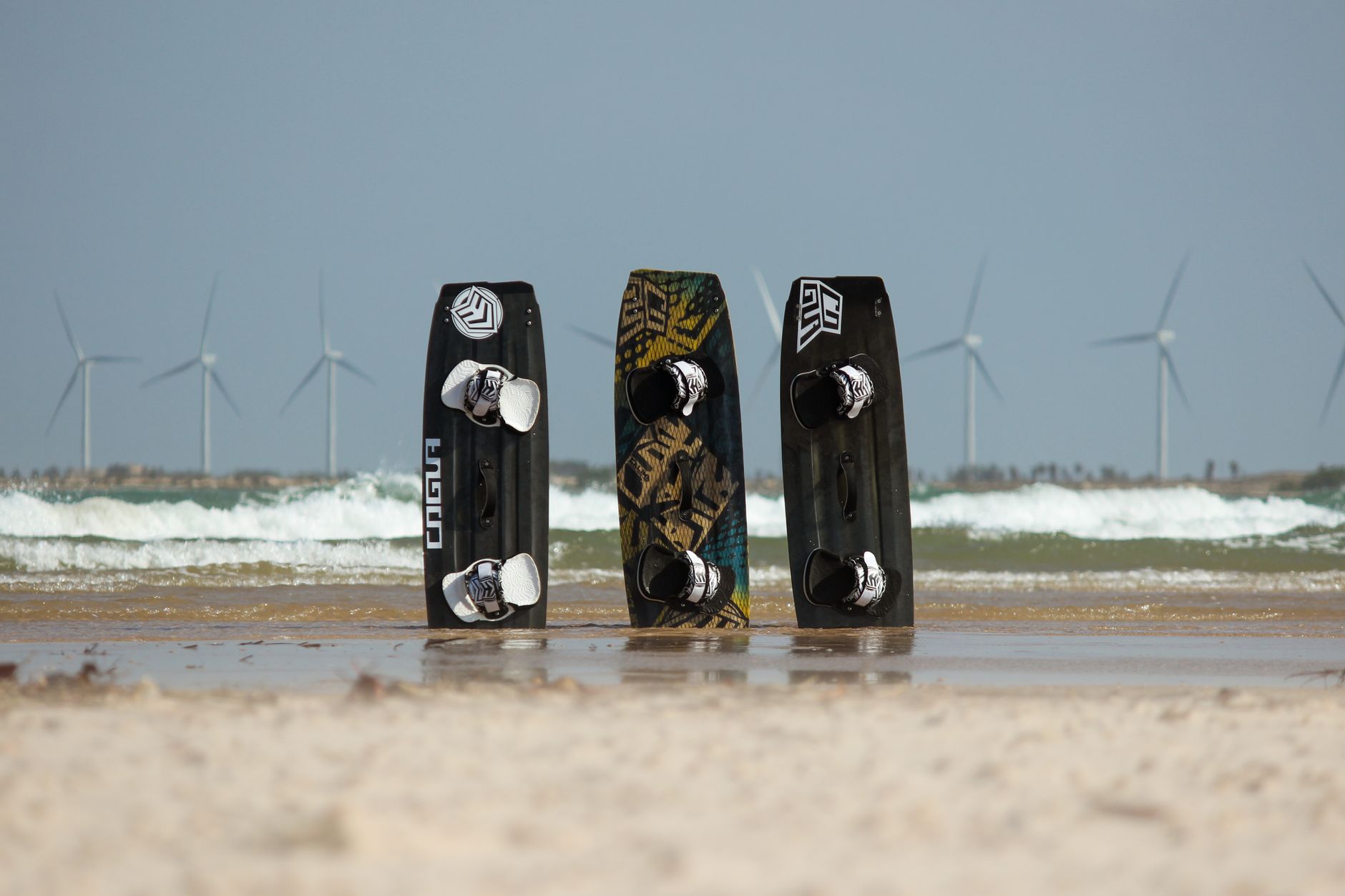 Three kiteboards on a sandy beach with wind turbines in Ceará, Brazil.