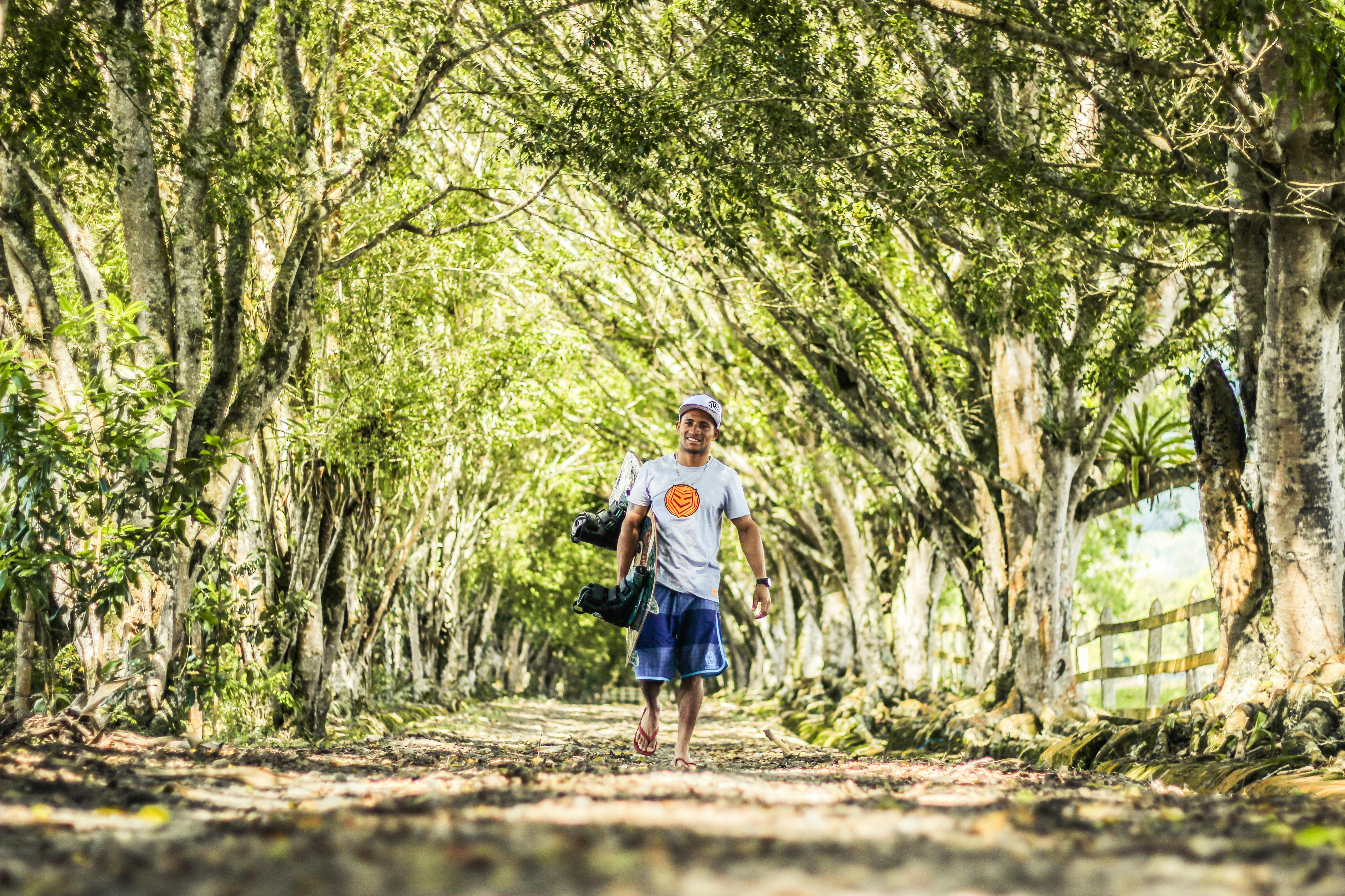 A man carrying a wakeboard walks along a sunlit tree-lined pathway in Colombia.