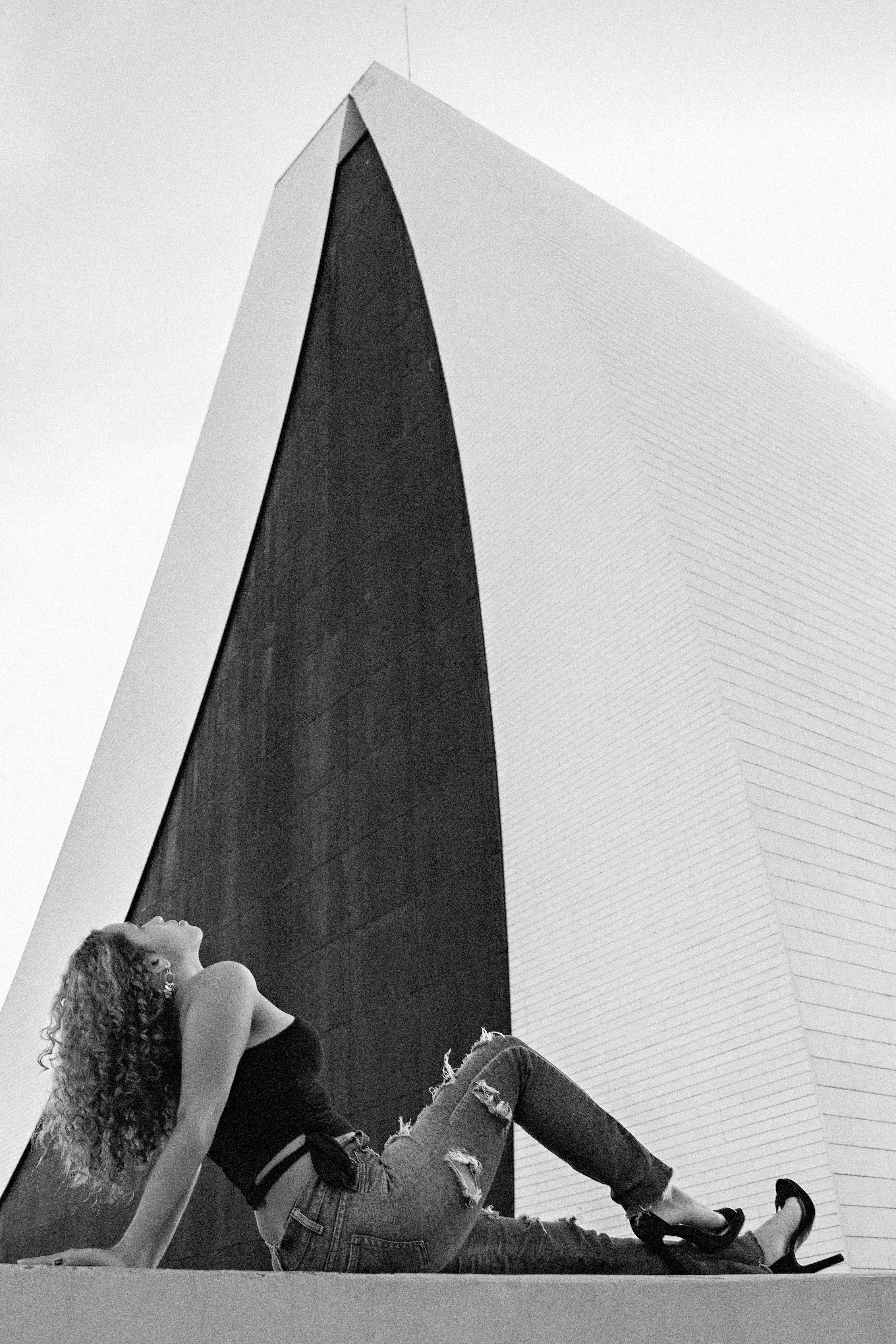 Black and white photo of a woman sitting by a modern architectural structure, capturing elegance and style.