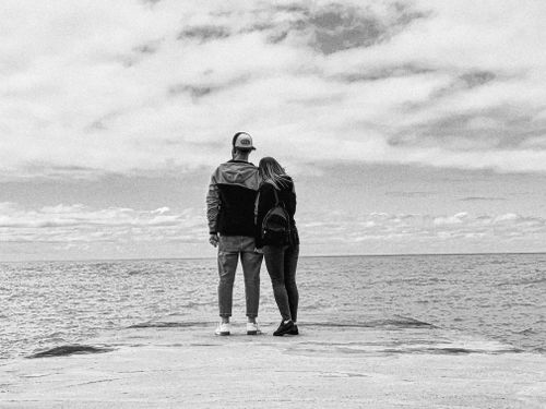 Free Black and white photo of a couple embracing on a pier with a seascape view under a cloudy sky. Stock Photo