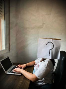 Woman sitting with a paper bag on head featuring a question mark, working on a laptop indoors.