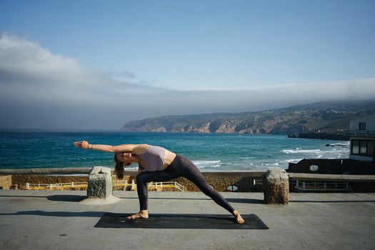 Woman performing yoga pose on a mat by the sea, with scenic coastal view.