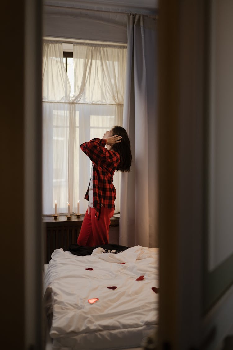 Woman In Her Pajamas Standing By Window In Bedroom