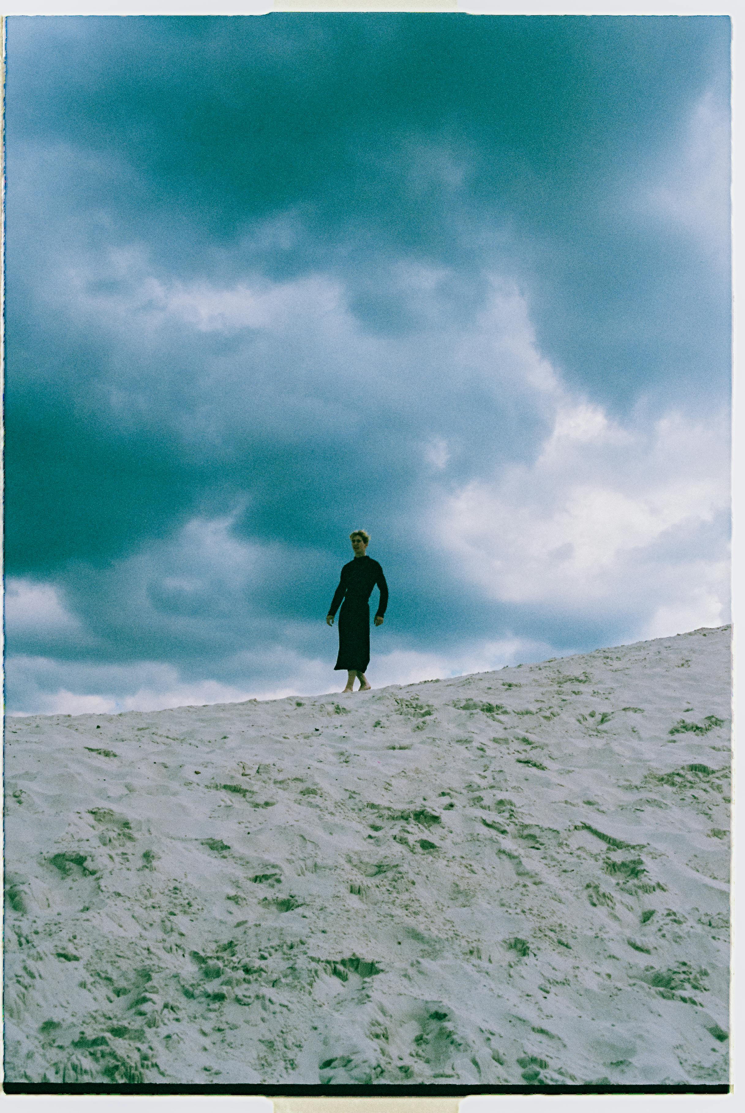A solitary figure in a dark dress stands on a sandy dune beneath a moody, cloud-filled sky.