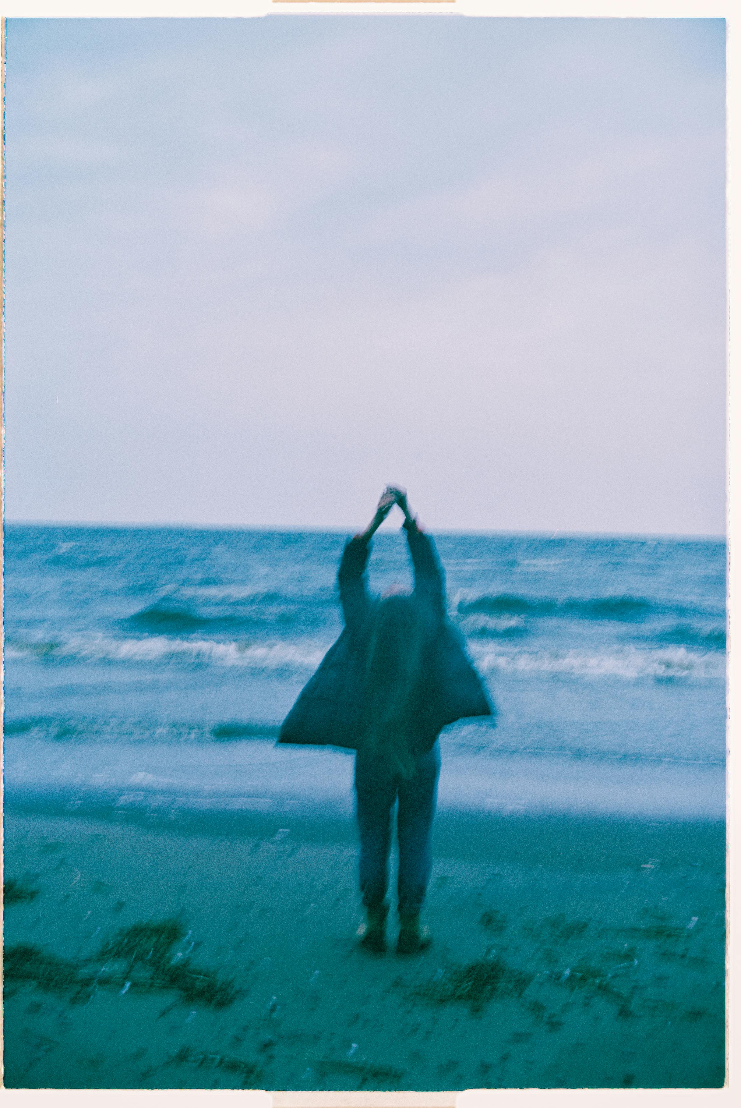A blurred silhouette of a person standing on a tranquil beach facing the sea during dusk.
