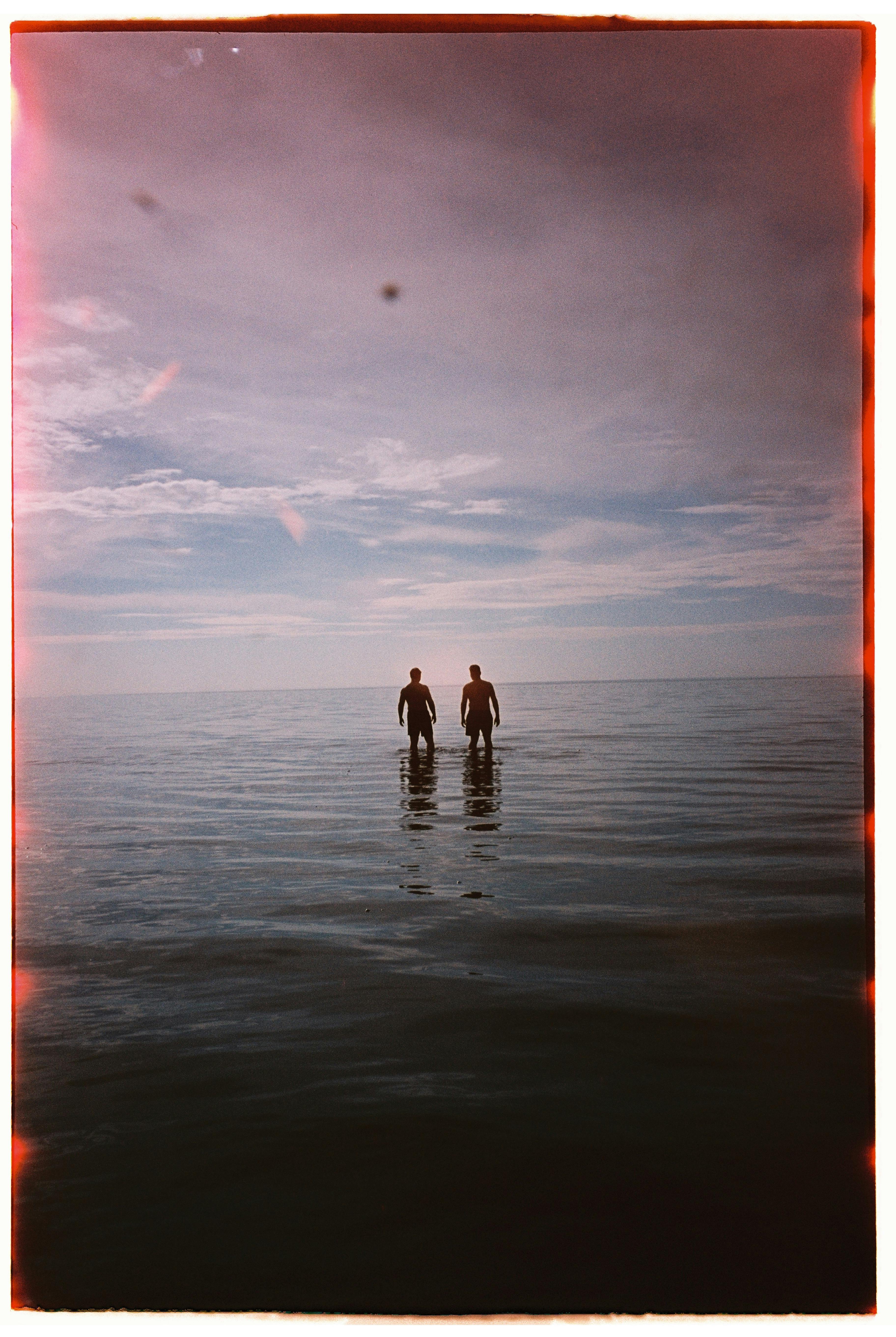 Two people wade through calm ocean waters, silhouetted against a wide sky, embodying tranquility and solitude.